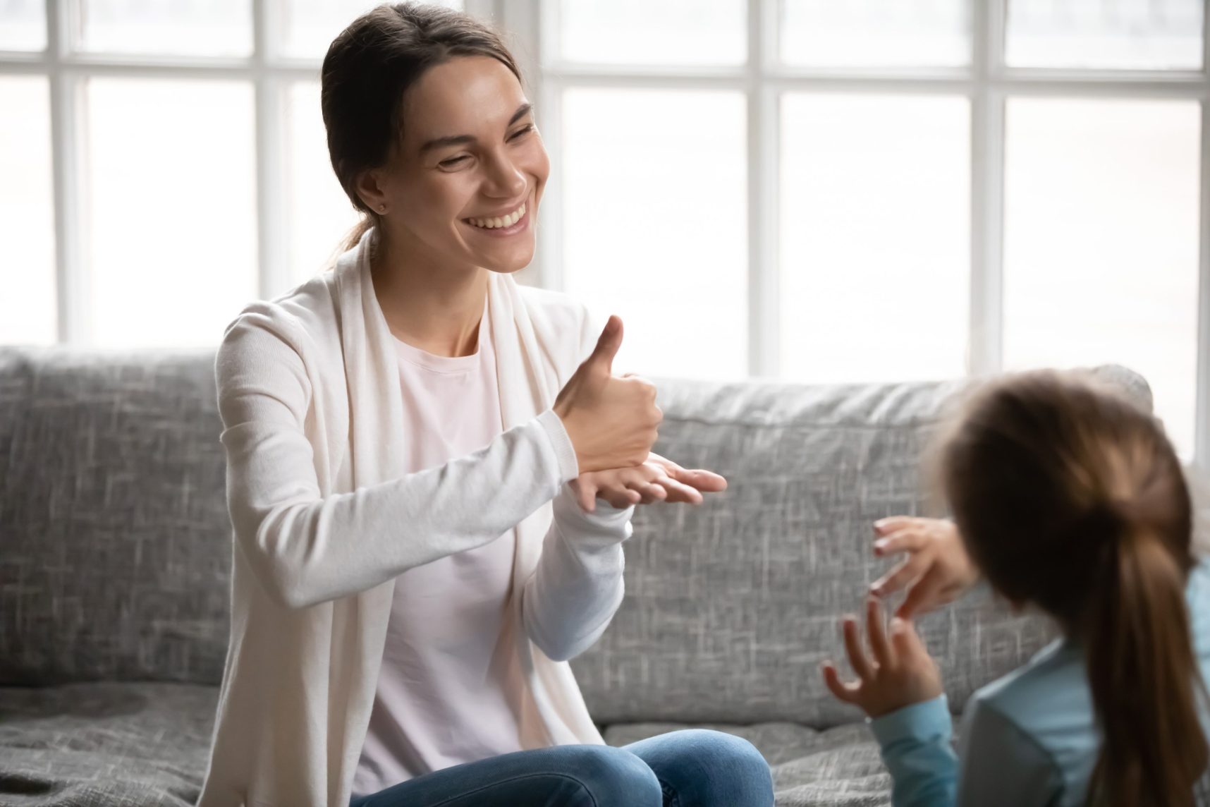 Smiling young Caucasian mom or nanny learn sign language with little disabled girl child at home. Happy positive teacher or tutor make hand gesture talk communicate nonverbal with small kid.