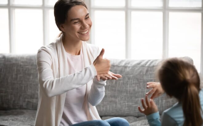 Smiling young Caucasian mom or nanny learn sign language with little disabled girl child at home. Happy positive teacher or tutor make hand gesture talk communicate nonverbal with small kid.