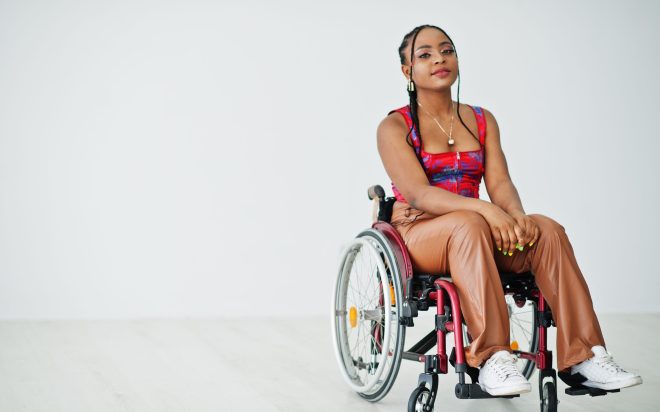 Young disabled African American woman in wheelchair against white wall.