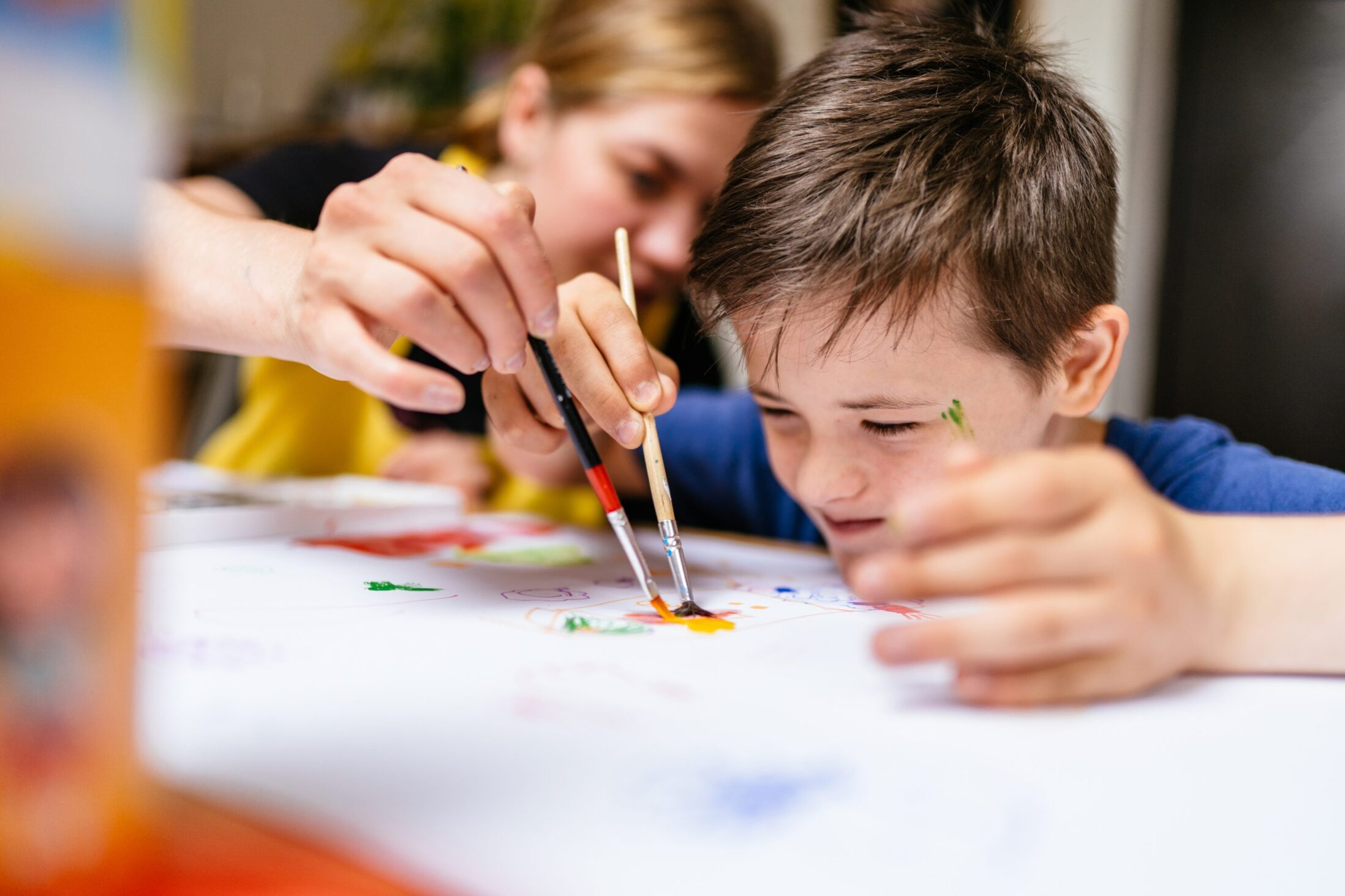 Young female teacher explain, talk at desk with a down syndrome schoolboy. Color painting for disabled kids, child who are down syndrome and student teacher, development of children with spesial needs