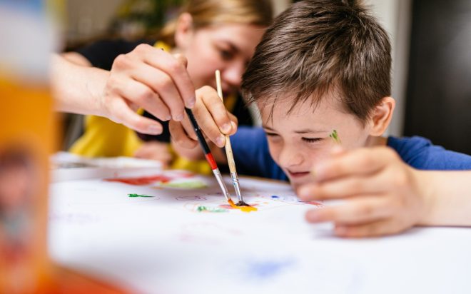 Young female teacher explain, talk at desk with a down syndrome schoolboy. Color painting for disabled kids, child who are down syndrome and student teacher, development of children with spesial needs