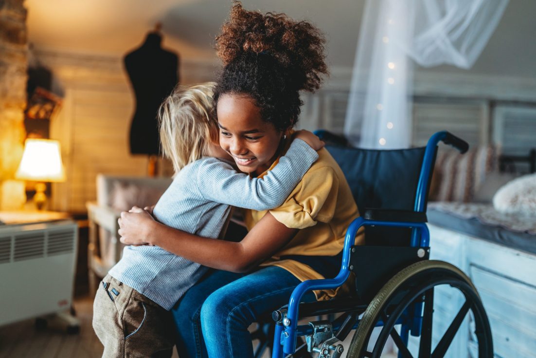 Happy multiethnic loving family. Smiling little girl with disability in wheelchair hugging with little brother at home